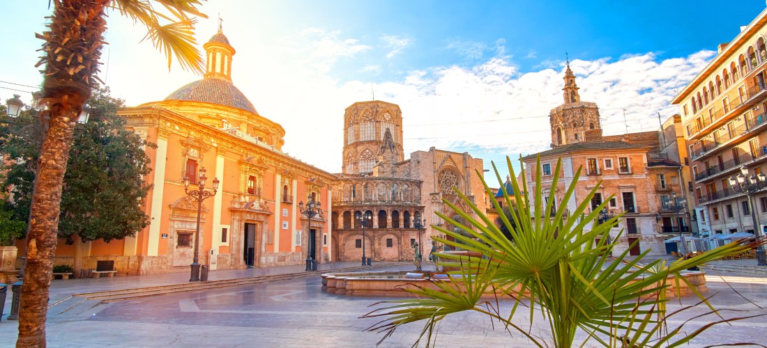 Blick auf einen sonnigen Platz in Valencia mit der barocken Basilika, gotischer Kathedrale und Palmen im Vordergrund. Historische Gebäude umrahmen die Szene.