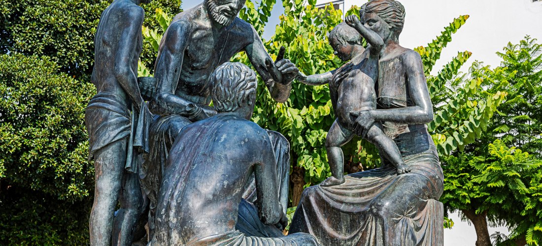 Bronzeskulpturengruppe, die den antiken griechischen Arzt Hippokrates zeigt, umgeben von Zuhörern und einer Frau mit Kind. Die Figuren sitzen und stehen in einer lebendigen Szene, als ob er medizinisches Wissen weitergibt. Im Hintergrund sind grüne Bäume 