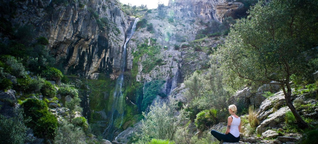 Frau sitzt auf Felsen vor Wasserfall und grüner Berglandschaft beim Wandern auf Teneriffa