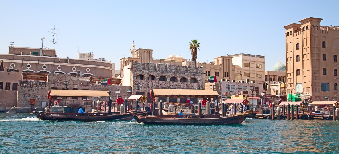 Traditionelle Holzdhausboote (Abras) auf dem Dubai Creek vor der historischen Altstadt mit sandfarbenen Gebäuden im arabischen Stil. Die Uferpromenade ist belebt mit Menschen, Märkten und kleinen Geschäften. Im Hintergrund sind Windtürme, eine blaue Mosch