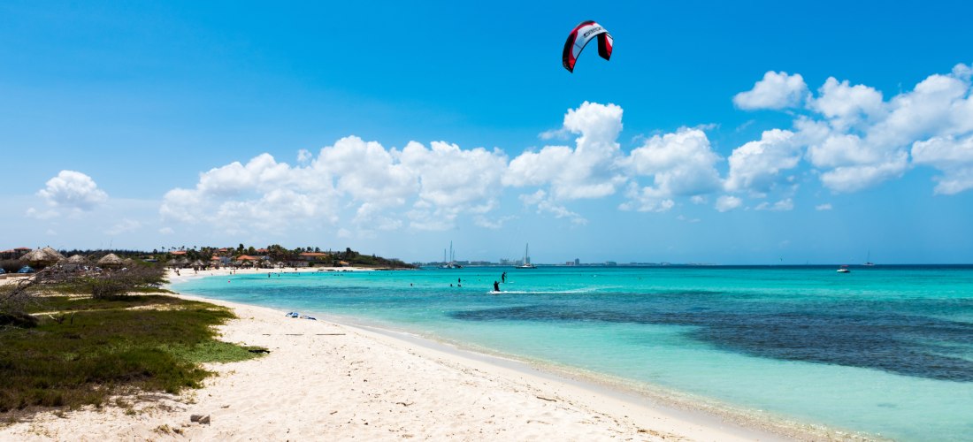 Blick über einen weißen Sandstrand auf Aruba mit türkisblauem Wasser. Ein Kitesurfer gleitet über die Wellen, der Drachen fliegt hoch am blauen Himmel. Im Hintergrund sind weitere Badegäste und Boote zu sehen.