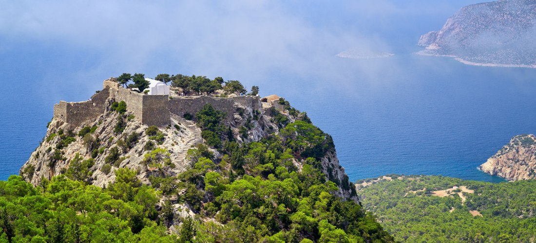 Blick auf eine mittelalterliche Festung mit weißer Kapelle, die spektakulär auf einem felsigen Bergrücken hoch über dem Meer thront. Eine steinerne Treppe führt durch grüne Pinienwälder hinauf zur Festung. Der Hintergrund zeigt das weite Blau des Meeres u