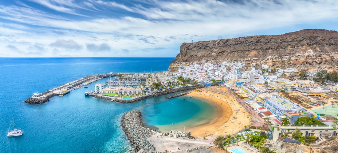 Luftaufnahme von Puerto de Mogán auf Gran Canaria mit Yachthafen, goldgelbem Strand und weißen Häusern vor einer Felskulisse.
