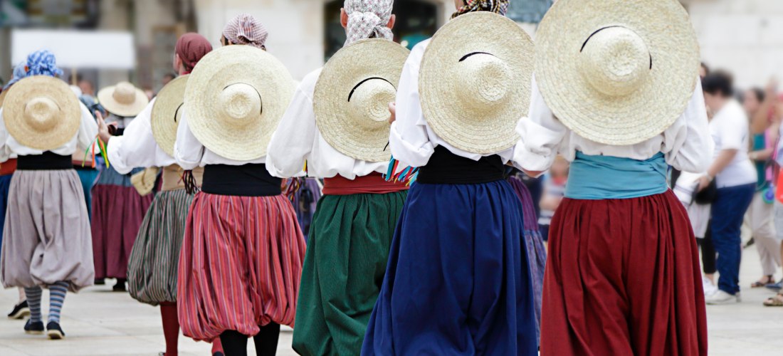 Gruppe von Menschen in traditioneller mallorquinischer Tracht, die auf einem öffentlichen Platz tanzen. Alle tragen weite bunte Röcke, weiße Blusen und Strohhüte auf dem Rücken. Die Szene spielt sich im Freien ab, umgeben von anderen Passanten.