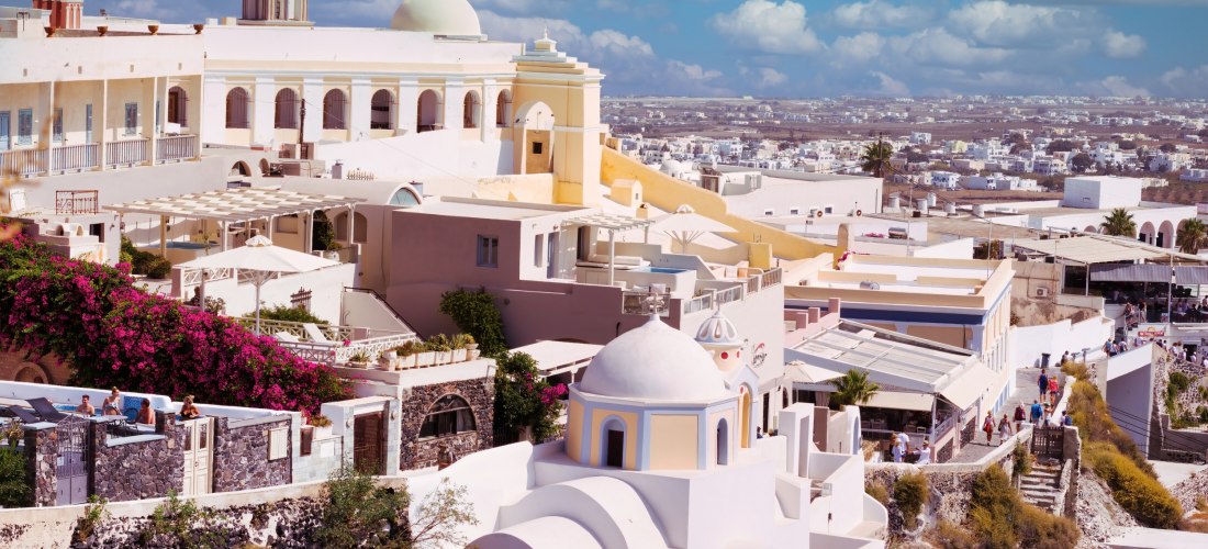 Panoramablick über die Stadt Fira auf der griechischen Insel Santorini. Weiße Gebäude mit Kuppeln und Glockentürmen erheben sich über steinige Hänge. Im Hintergrund ist die weite Landschaft der Insel mit verstreuten Häusern zu sehen, unter einem Himmel mi