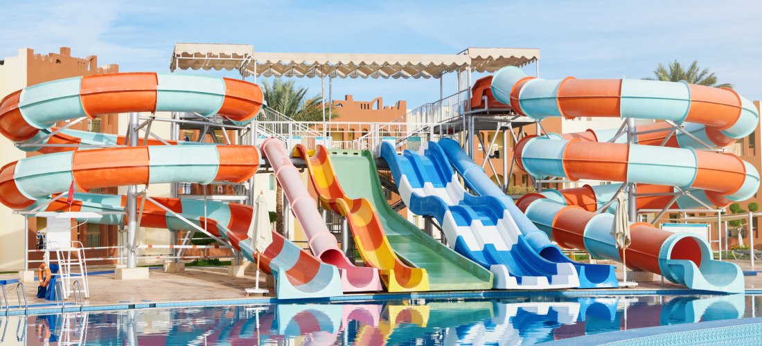 Großer Wasserpark mit bunten Wasserrutschen, die in einen klaren Pool führen. Im Hintergrund stehen Hotelgebäude, der Himmel ist blau und wolkenlos.