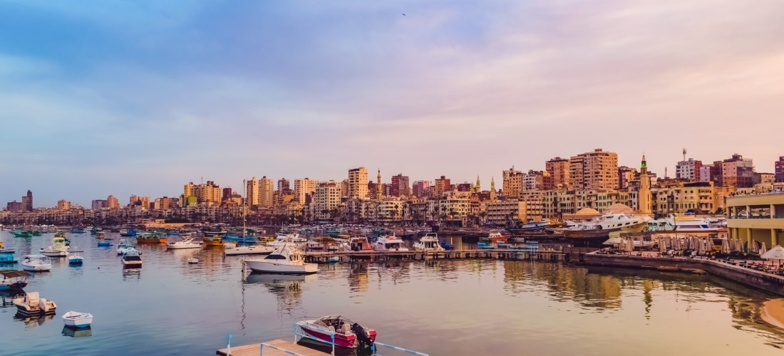 Ägypten - Alexandria Das Bild zeigt den Hafen von Alexandria in Ägypten in der Abenddämmerung. Im ruhigen Wasser liegen zahlreiche bunte Boote, kleinere Yachten und Fischerboote vor Anker. Die Stadt erstreckt sich im Hintergrund mit einer dichten Skyline aus mehrstöckigen Woh