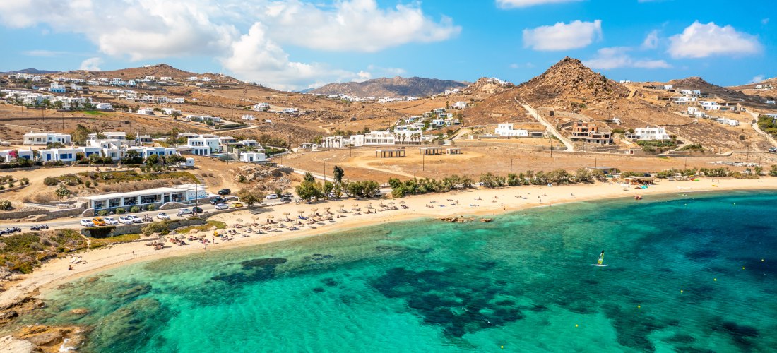Luftaufnahme vom Kalafatis Beach auf Mykonos mit goldenem Sandstrand, Sonnenschirmen und klarem türkisblauem Wasser