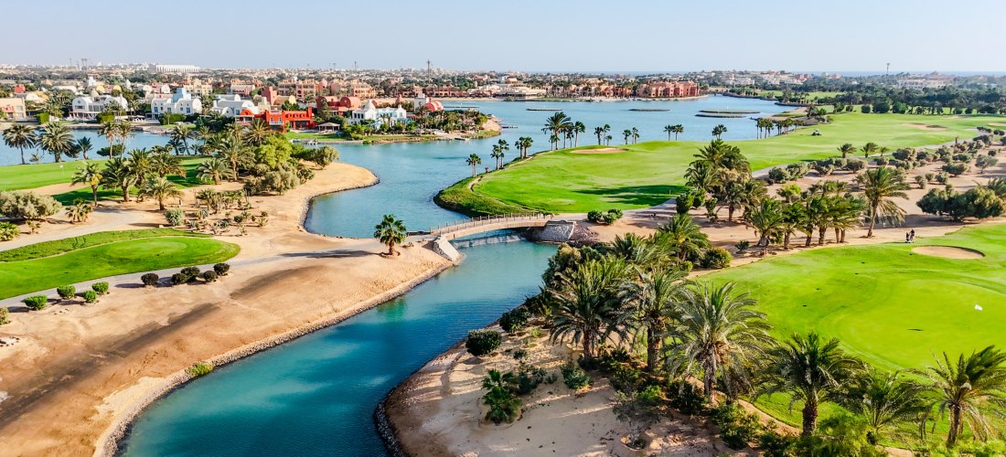 Blick auf eine gepflegte Golfanlage in El Gouna, Ägypten. Zwischen sattem Grün und Palmen verlaufen künstliche Wasserkanäle. Im Hintergrund sind bunte Villen und Häuser entlang der Lagunen zu sehen, mit der Stadt El Gouna in weiter Ferne unter blauem Himm