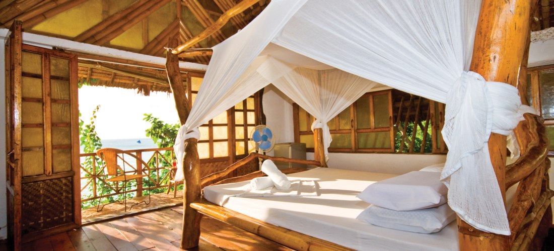 Offenes Schlafzimmer mit Bambusmöbeln und Himmelbett mit Moskitonetz. Der Raum hat Holzboden, eine hohe Decke und Blick auf das Meer durch geöffnete Türen zum Balkon.