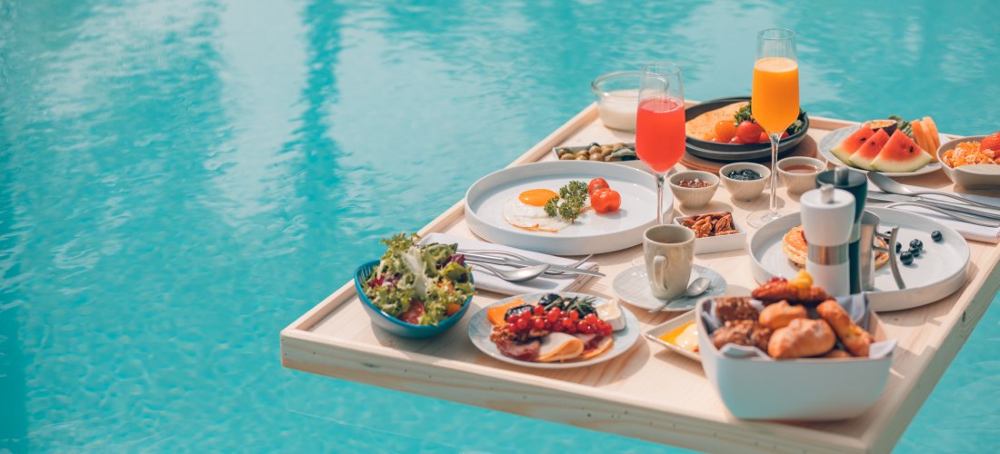 Schwimmendes Frühstückstablett mit Eiern, Obst, Gebäck und Getränken auf einem Pool