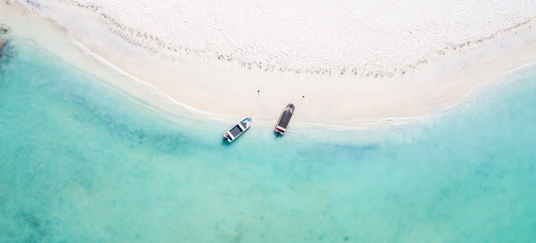 Zwei kleine Boote liegen im seichten, türkisfarbenen Wasser an einem weißen Sandstrand. Die Szene ist aus der Vogelperspektive aufgenommen, der Strand wirkt ruhig und unberührt.