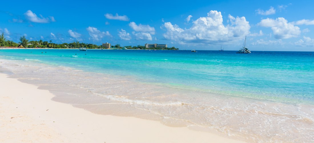 Weißer Sandstrand mit türkisblauem Meer, Palmen und Booten im Hintergrund auf Barbados.