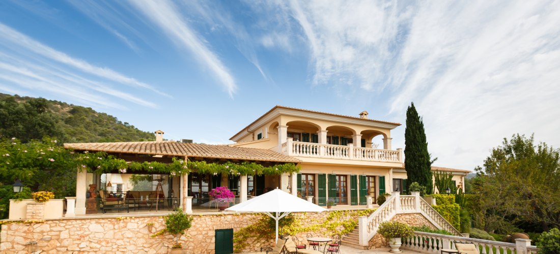 Große mediterrane Villa mit mehreren Stockwerken, großzügigem Balkon und überdachter Terrasse. Vor dem Haus befindet sich ein Pool mit Liegestühlen und Sonnenschirm, umgeben von Steinmauern und Zierpflanzen. Der Himmel ist blau mit leichten Schleierwolken