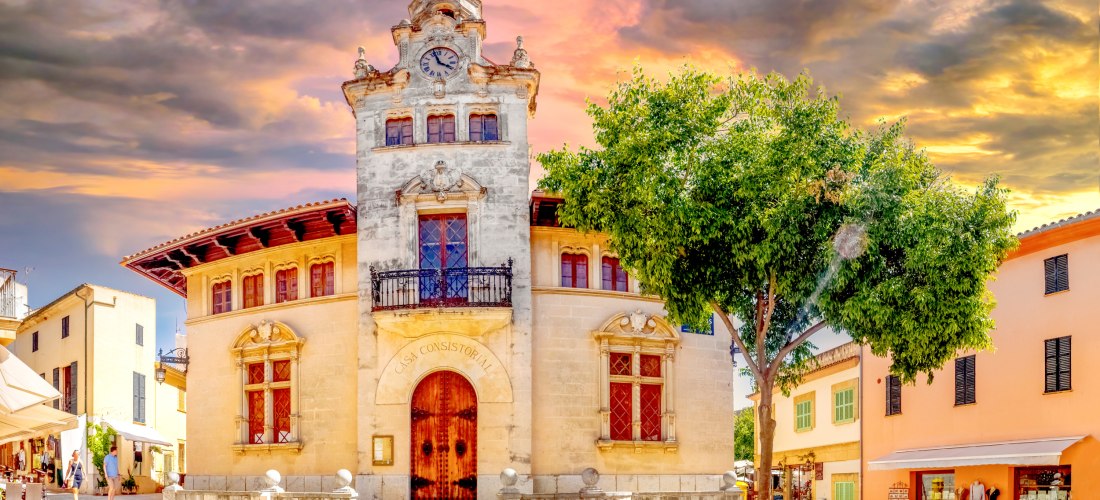 Blick auf das historische Rathaus Casa Consistorial in Alcúdia Altstadt auf Mallorca. Das Gebäude ist aus hellem Naturstein erbaut und hat eine dekorative Fassade mit geschnitzten Balkonen, Fensterläden und einem markanten Uhrturm mit Zinnen. Davor führt 