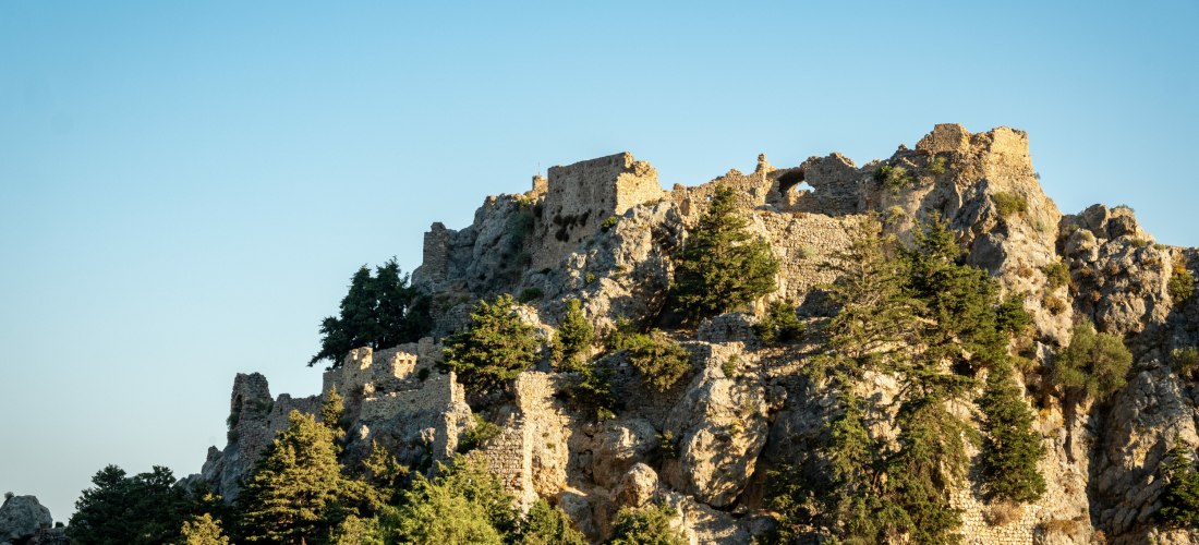 Mittelalterliche Burgruine auf steilem Felsen, umgeben von Pinienbäumen unter klarem Himmel im Sonnenlicht