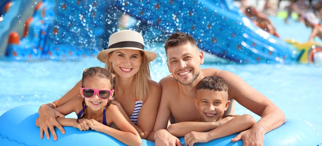 Fröhliche Familie mit zwei Kindern auf einem Schwimmreifen im Wasserpark, im Hintergrund spritzt Wasser von einer blauen Rutsche