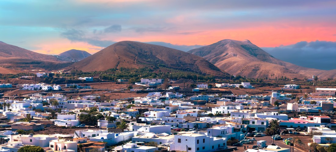 Blick auf das Dorf Yaiza auf Lanzarote mit weißen Häusern, Vulkanlandschaft und Bergen im Hintergrund bei Sonnenuntergang.