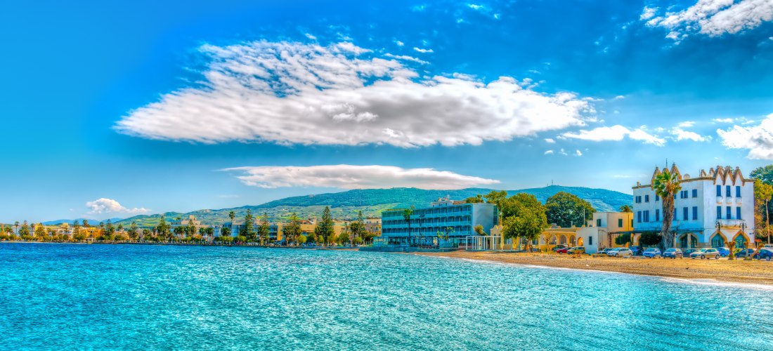 Blick auf die Uferpromenade von Kos-Stadt bei Sonnenschein. Im Vordergrund glitzert das türkisfarbene Wasser der Ägäis. Am Ufer stehen mehrere Gebäude, darunter ein modernes Hotel mit Balkonen und ein weißes, historisch wirkendes Gebäude mit Palmen davor.