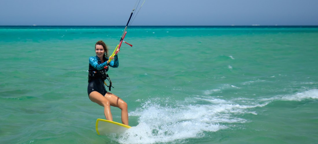 Eine junge Frau fährt lachend auf einem gelben Kiteboard über das türkisfarbene Meer. Sie hält sich an den Lenkstangen des Kites fest, während Wasser hinter ihr aufspritzt. Der Horizont ist klar, der Himmel wolkenlos – ein perfekter Moment voller Action, 
