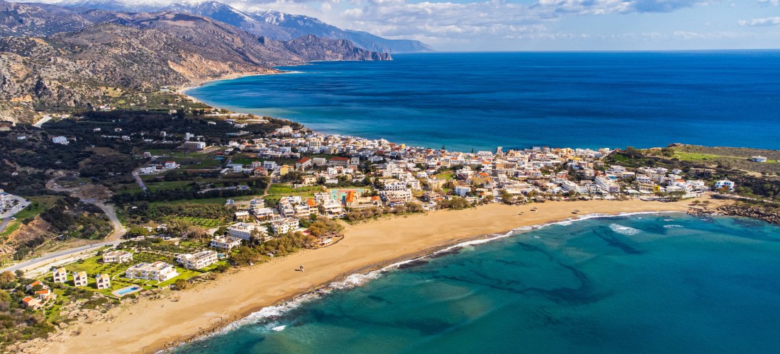 Luftaufnahme eines Küstenortes auf Kreta mit Sandstrand, blauem Meer und weißen Häusern entlang der Küste