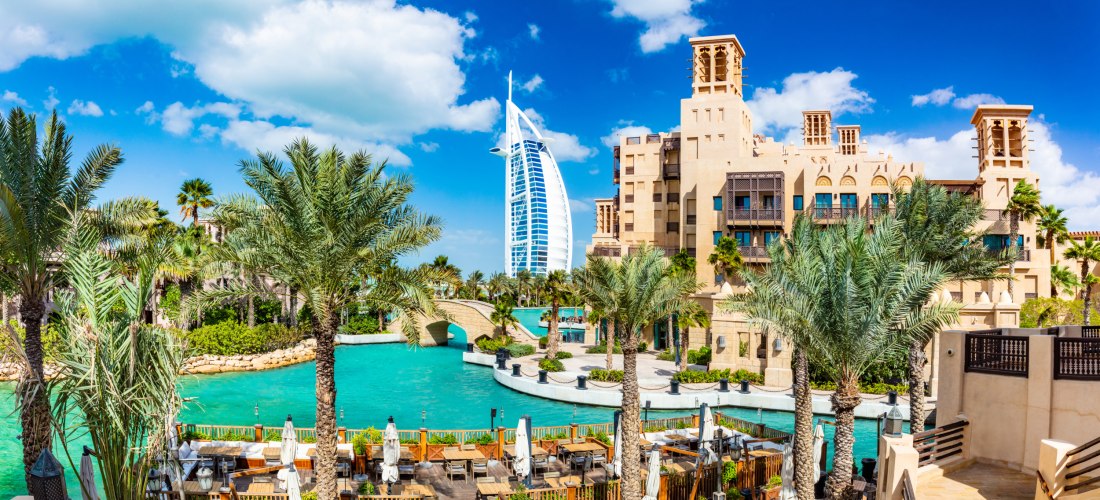 Dubai - Jumeirah Panoramaaufnahme eines luxuriösen Resorts in Dubai mit Wasserkanälen, Palmen und traditionellen Gebäuden im arabischen Stil. Im Hintergrund ragt das berühmte Hotel Burj Al Arab mit seiner segelförmigen Architektur in den Himmel. Im Vordergrund befinden si