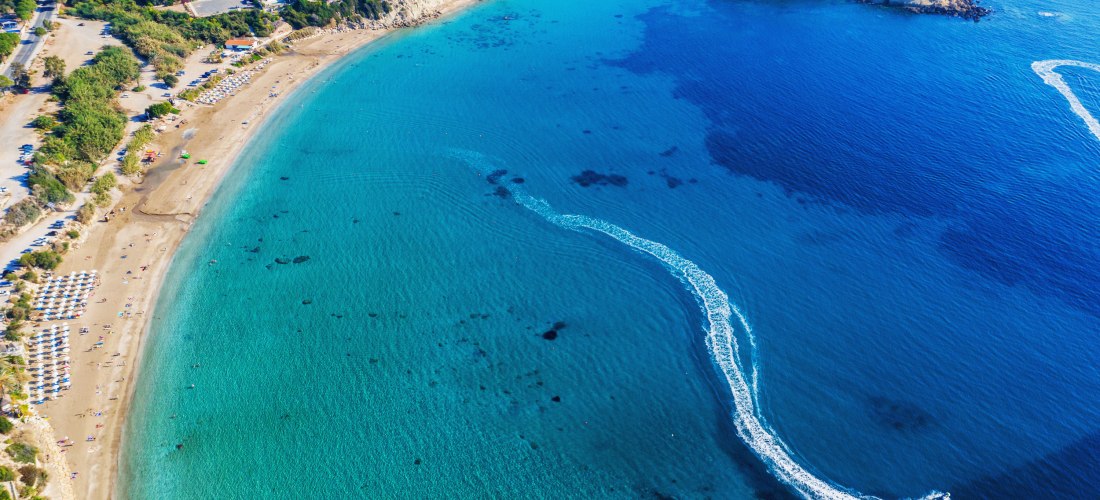 Luftaufnahme einer weiten Sandbucht auf Zypern mit türkisblauem Wasser, Klippen und der Spur eines Motorbootes im Meer.