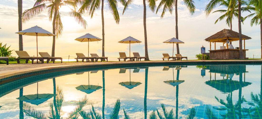 Luxuriöser Poolbereich mit Liegestühlen, Sonnenschirmen und Palmen, die sich im ruhigen Wasser spiegeln, bei Sonnenuntergang am Strand