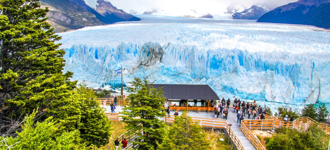Eine große Besucherplattform mit Holzgeländern vor dem gewaltigen Perito-Moreno-Gletscher in Island. Im Hintergrund sind schneebedeckte Berge und dramatische Wolkenformationen zu sehen. Menschen stehen auf der Plattform und bestaunen das beeindruckende Na