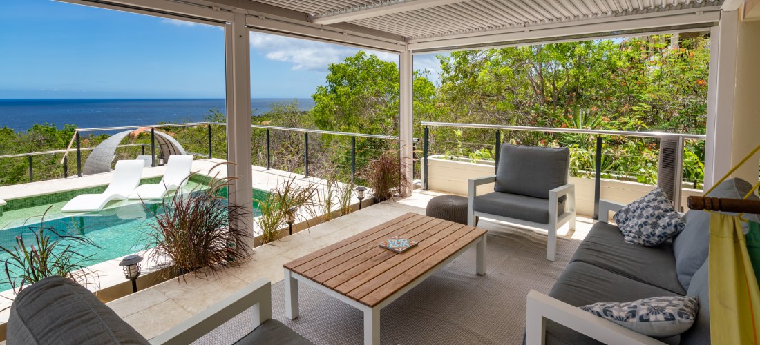 Überdachte Terrasse mit gemütlichen Sitzmöbeln und Blick auf einen privaten Pool mit zwei weißen Liegen im Wasser. Dahinter dichter grüner Bewuchs und ein weiter Blick aufs tiefblaue Meer unter klarem Himmel. Eine entspannte, tropische Atmosphäre.