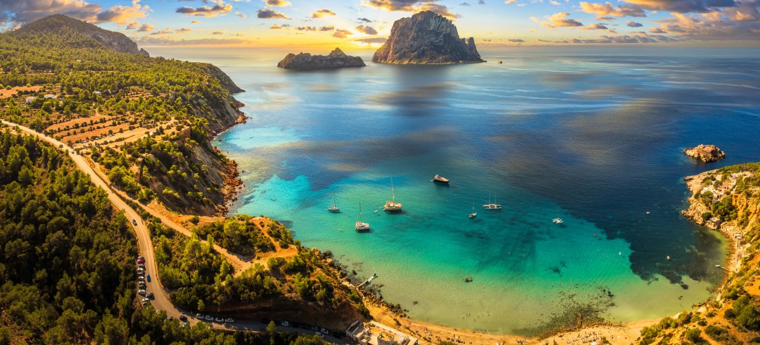 Blick von der Küste bei Cala d’Hort auf die unbewohnte Felseninsel Es Vedrà vor Ibiza. Türkisfarbenes Meer, mehrere Segelboote im Wasser und ein strahlender Himmel mit Wolken bei Sonnenuntergang.