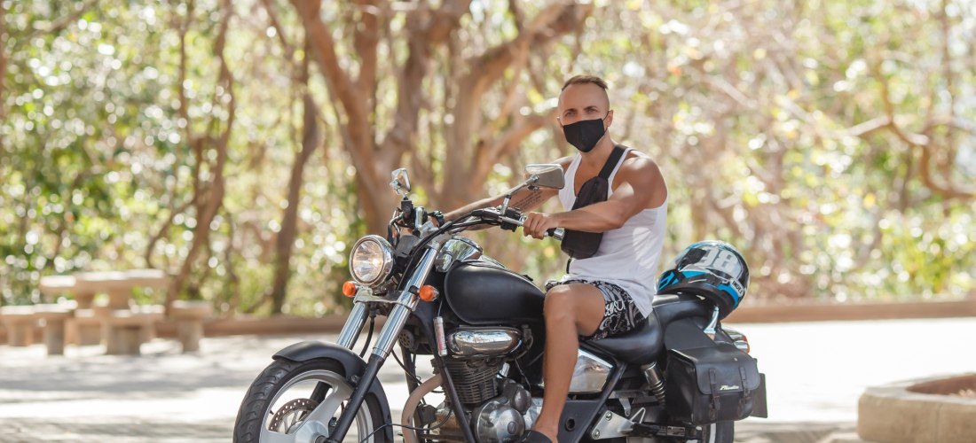 Ein Mann mit kurzer Frisur sitzt auf einem schwarzen Motorrad. Er trägt ein weißes Tanktop, Shorts, schwarze Sandalen und eine schwarze Gesichtsmaske. Neben ihm auf dem Sitz liegt ein Helm. Im Hintergrund sind Bäume mit hellem Sonnenlicht zu sehen.