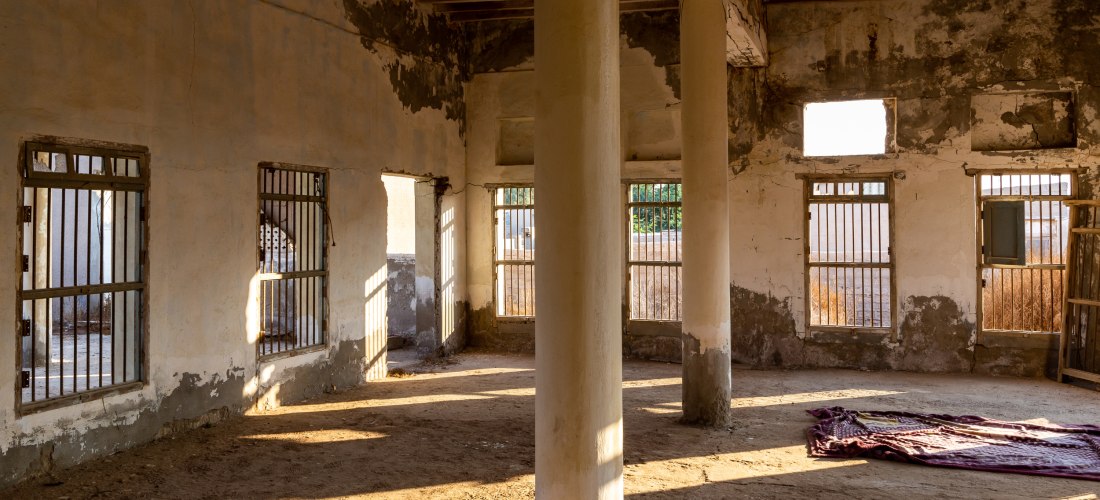 Innenansicht eines verlassenen Gebäudes in Al Jazirah Al Hamra mit Sonnenlicht, vergitterten Fenstern und historischen Mauern.