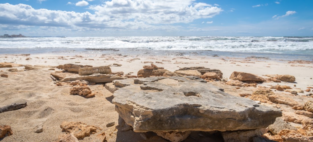 Felsiger Sandstrand mit flachen Steinen im Vordergrund und Wellen im Hintergrund unter teils bewölktem Himmel
