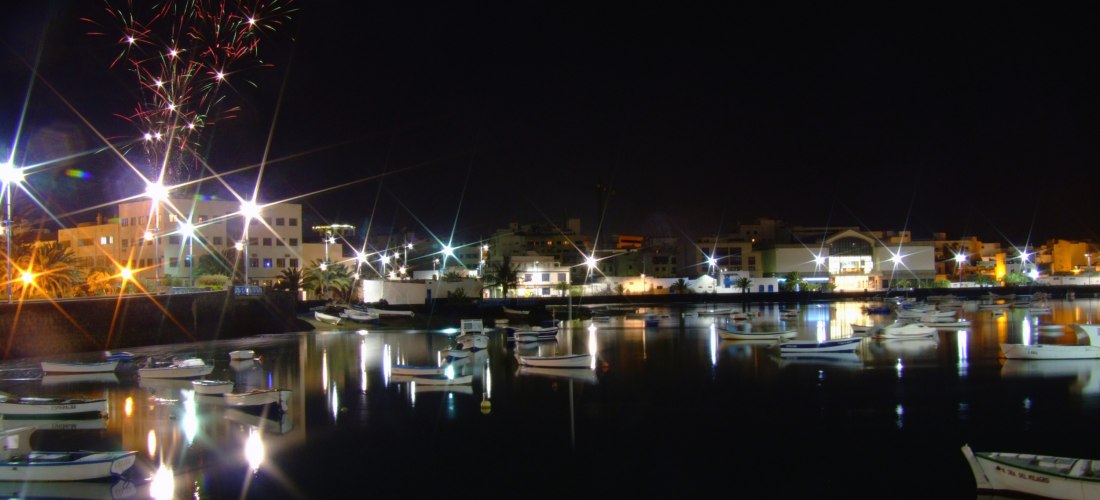 Nächtlicher Hafen mit kleinen Booten, beleuchtet von Laternen. Im Hintergrund erhellen bunte Feuerwerkskörper den Himmel über den Häusern der Stadt.