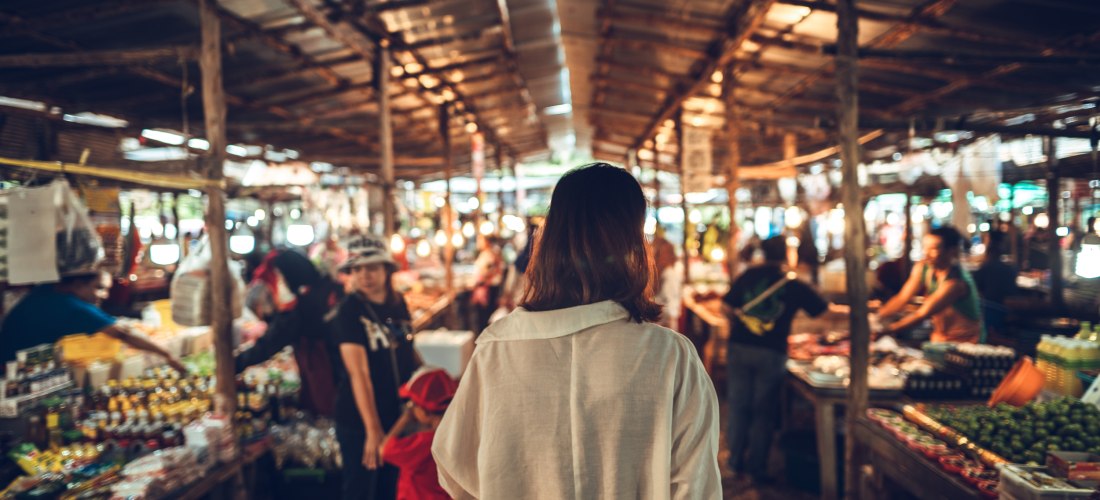 Das Bild zeigt eine Frau mit schulterlangem Haar und weißem Hemd von hinten, wie sie durch einen traditionellen Markt in Südostasien läuft. Der Markt ist überdacht mit Wellblechdächern und von warmem Licht durchzogen. Links und rechts reihen sich Verkaufs