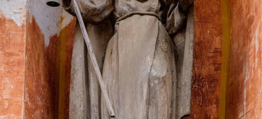 Steinerne Statue eines Heiligen in einer Wandnische mit rot-orange Patina. Die Figur trägt ein langes Gewand, hält in einer Hand einen Stab mit einer Glocke und in der anderen ein aufgeschlagenes Buch. Zu Füßen der Statue liegt ein kleines steinernes Schw