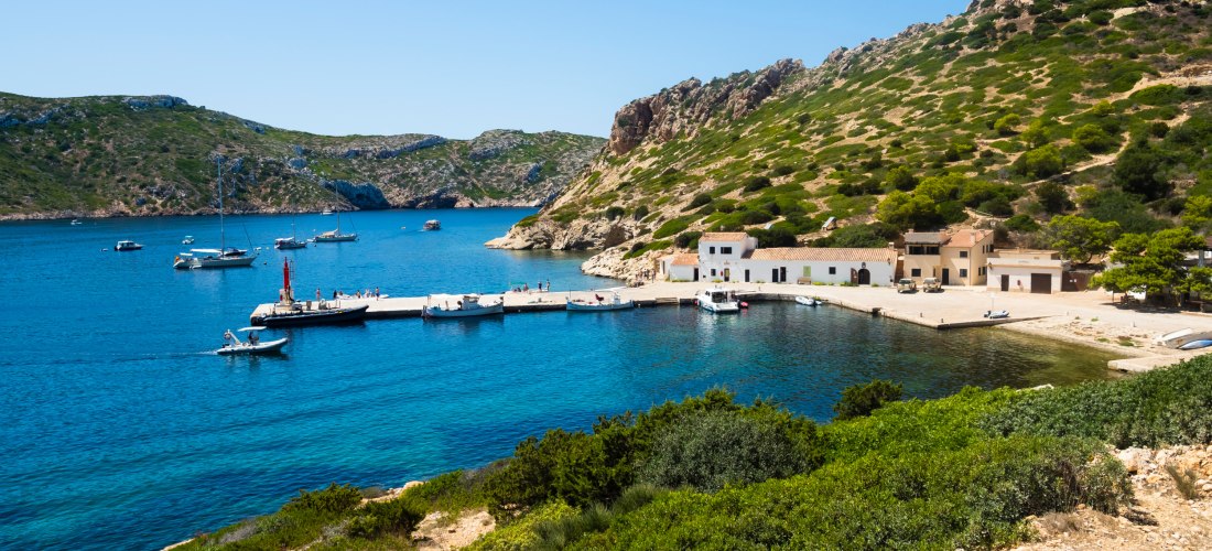 Eine kleine, idyllische Hafenbucht mit türkisblauem Wasser, umgeben von felsigen Hügeln, die mit niedrigen Büschen und Sträuchern bewachsen sind. Am Ufer stehen weiße Häuser mit roten Ziegeldächern und kleinen Booten vor der Mole. Auf einem Hügel thront e