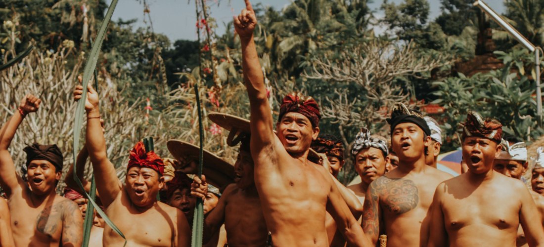 Eine Gruppe balinesischer Männer in traditioneller Kleidung steht versammelt unter freiem Himmel. Viele tragen Sarongs, einige heben die Arme und rufen oder jubeln. Einer der Männer zeigt mit ausgestrecktem Finger nach oben. Die Szene wirkt lebendig und r