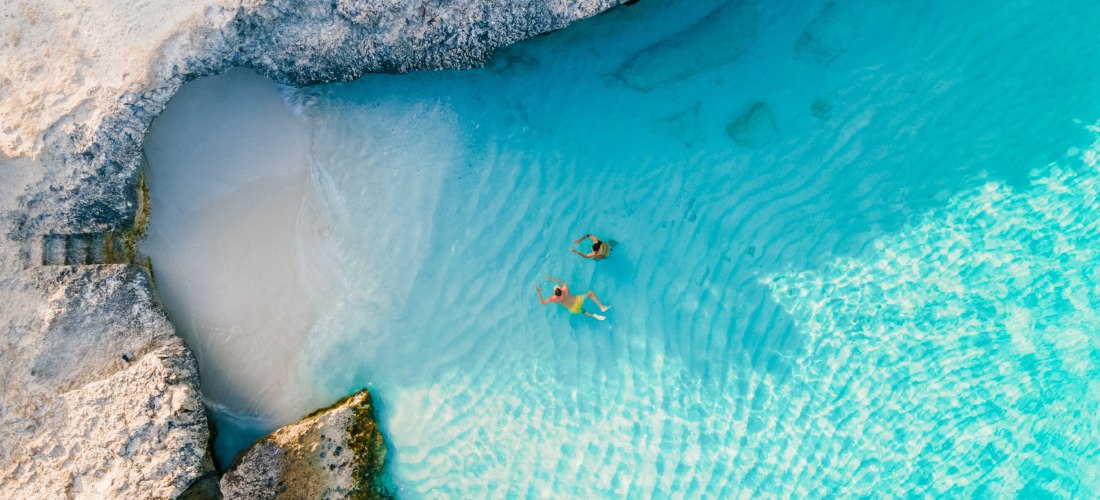 Zwei Menschen schwimmen in glasklarem, türkisblauem Wasser einer Bucht auf Aruba. Umgeben von hellen Felsen und Sand, eingefangen aus der Vogelperspektive.