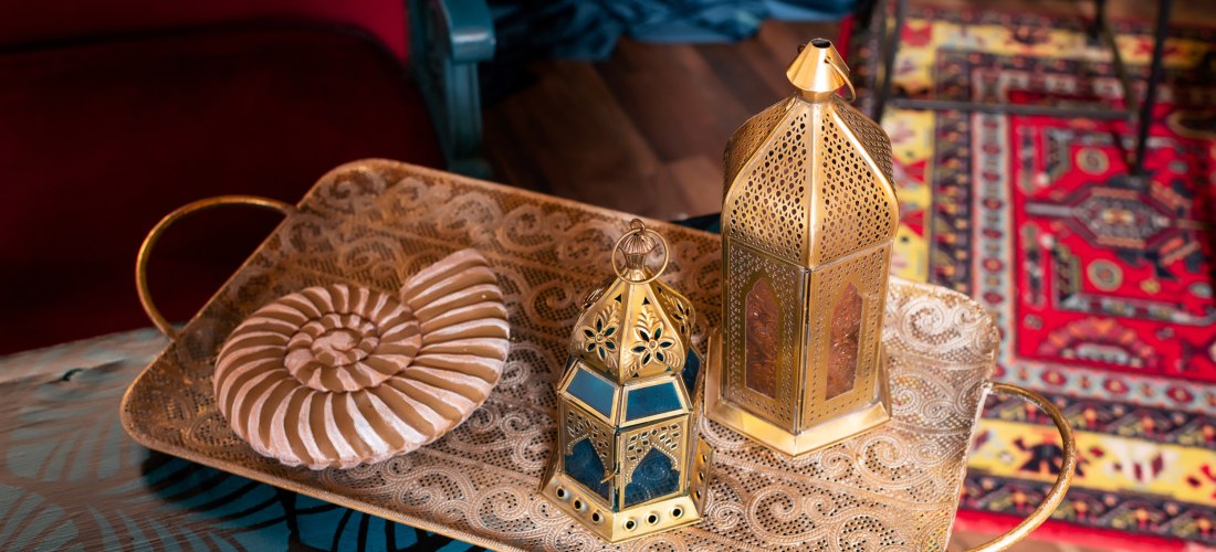 Ein kunstvoll verziertes goldenes Tablett mit zwei traditionellen orientalischen Laternen und einer dekorativen Muschel in Spiralenform steht auf einem bunt gemusterten Tisch. Im Hintergrund sind ein roter Teppich mit orientalischem Muster und ein Samtvor