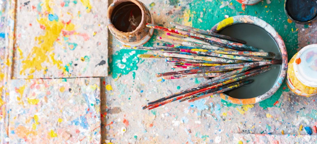 Mehrere bunte, gebrauchte Pinsel stehen in einem Farbtopf auf einem mit Farbspritzern bedeckten Boden. Umgeben sind sie von offenen Farbdosen in kräftigen Farben wie Gelb und Schwarz.