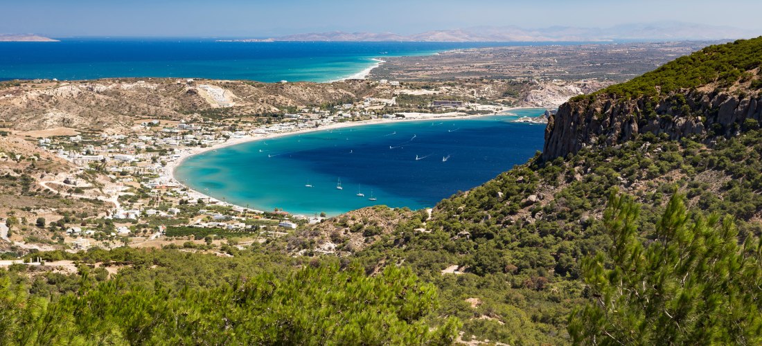 Weitläufiger Blick über die Kefalos-Bucht im Südwesten der Insel Kos. Die halbmondförmige Bucht zeigt türkis- bis dunkelblaues Wasser mit zahlreichen kleinen Segelbooten. Rund um die Bucht liegen weiße Häuser, trockene Hänge und grüne Kiefernwälder.