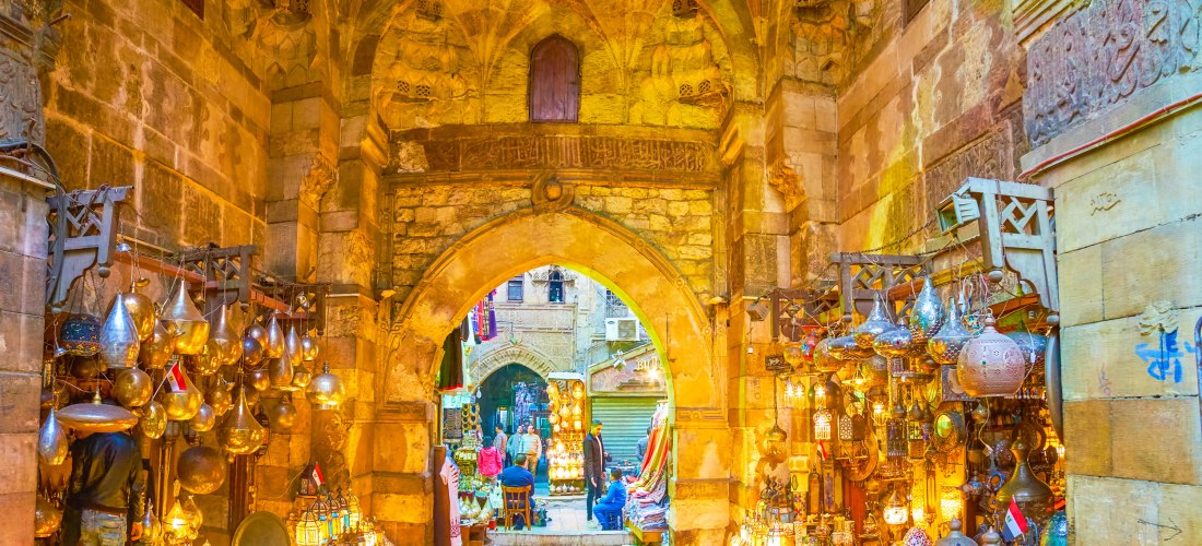 Ägypten - Kairo Das Bild zeigt den farbenfrohen Eingang zum Khan el-Khalili-Basar in Kairo, einem der bekanntesten Märkte Ägyptens. Die Szene spielt sich unter einem historischen Steingewölbe mit arabischer Architektur ab. Links und rechts des Durchgangs stapeln sich kun