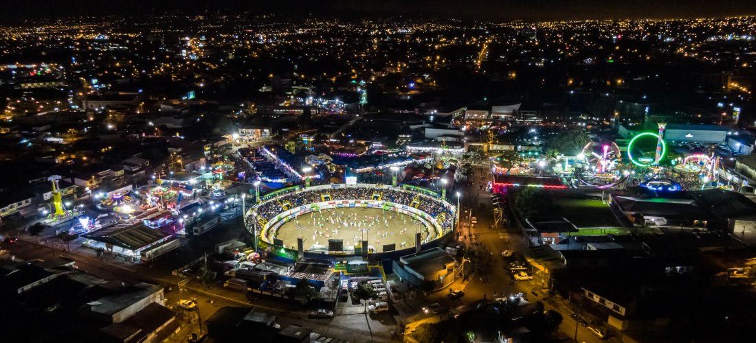 Luftaufnahme eines beleuchteten Festgeländes bei Nacht mit einer großen, runden Arena im Zentrum und zahlreichen bunten Fahrgeschäften drumherum.