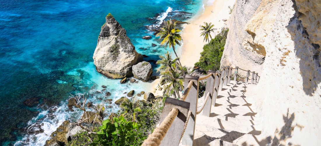 Steile Treppe mit Seilgeländer führt an einer Felswand hinunter zu einem traumhaften Strand mit türkisblauem Wasser und Palmen