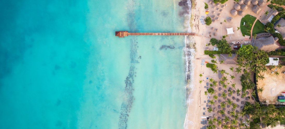 Drohnenaufnahme eines tropischen Strandes mit Holzsteg im türkisblauen Meer, Palmen und Liegestühlen entlang des Sandstrandes