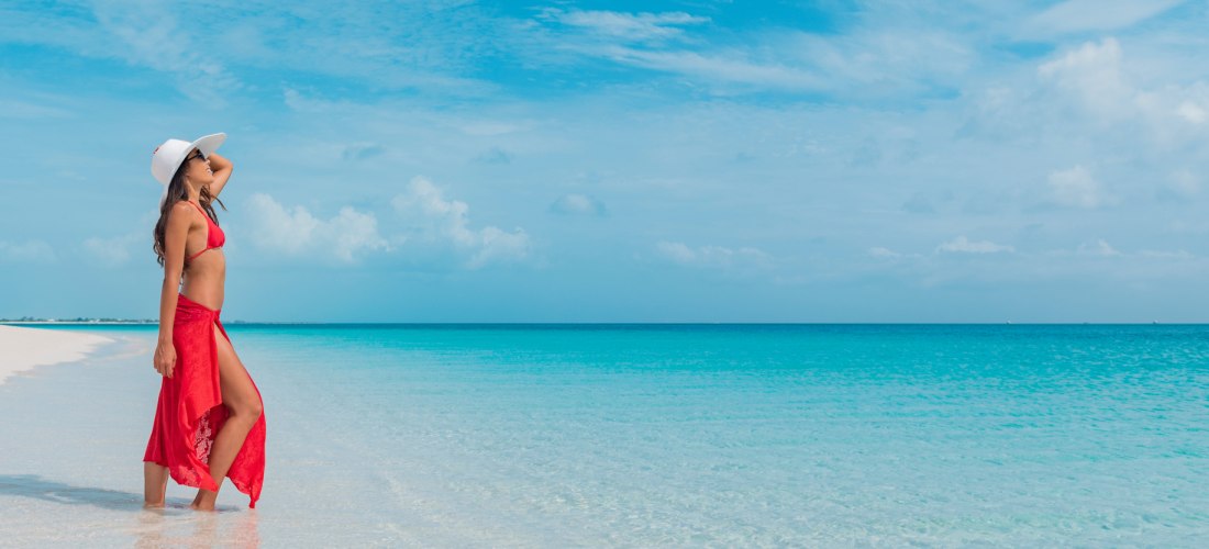 Frau in rotem Bikini und weißem Sonnenhut steht entspannt am weißen Sandstrand mit türkisfarbenem Wasser unter einem blauen Himmel
