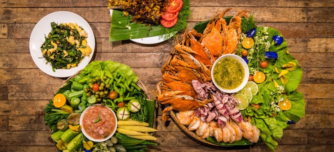 Das Bild zeigt eine bunte Auswahl traditioneller thailändischer Speisen, angerichtet auf einem rustikalen Holztisch. Rechts befindet sich eine große Platte mit gekochten Meeresfrüchten – darunter Garnelen, Tintenfisch und Krabben – garniert mit Limetten, 