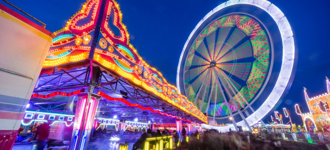 Bunte Kirmesszene bei Nacht mit einem leuchtenden Riesenrad im Hintergrund und einem hell erleuchteten Autoscooter im Vordergrund. Viele Besucher schlendern durch das Gelände. Die bunten Lichter erzeugen Bewegungsspuren, die die dynamische Atmosphäre des 